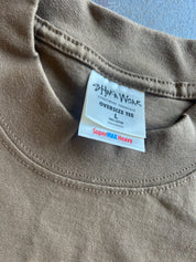 Close-up of a taupe brown oversized cotton t-shirt showing the inner neck label which reads 'SHAWA WEAR' and 'SuperMAX Heavy'.