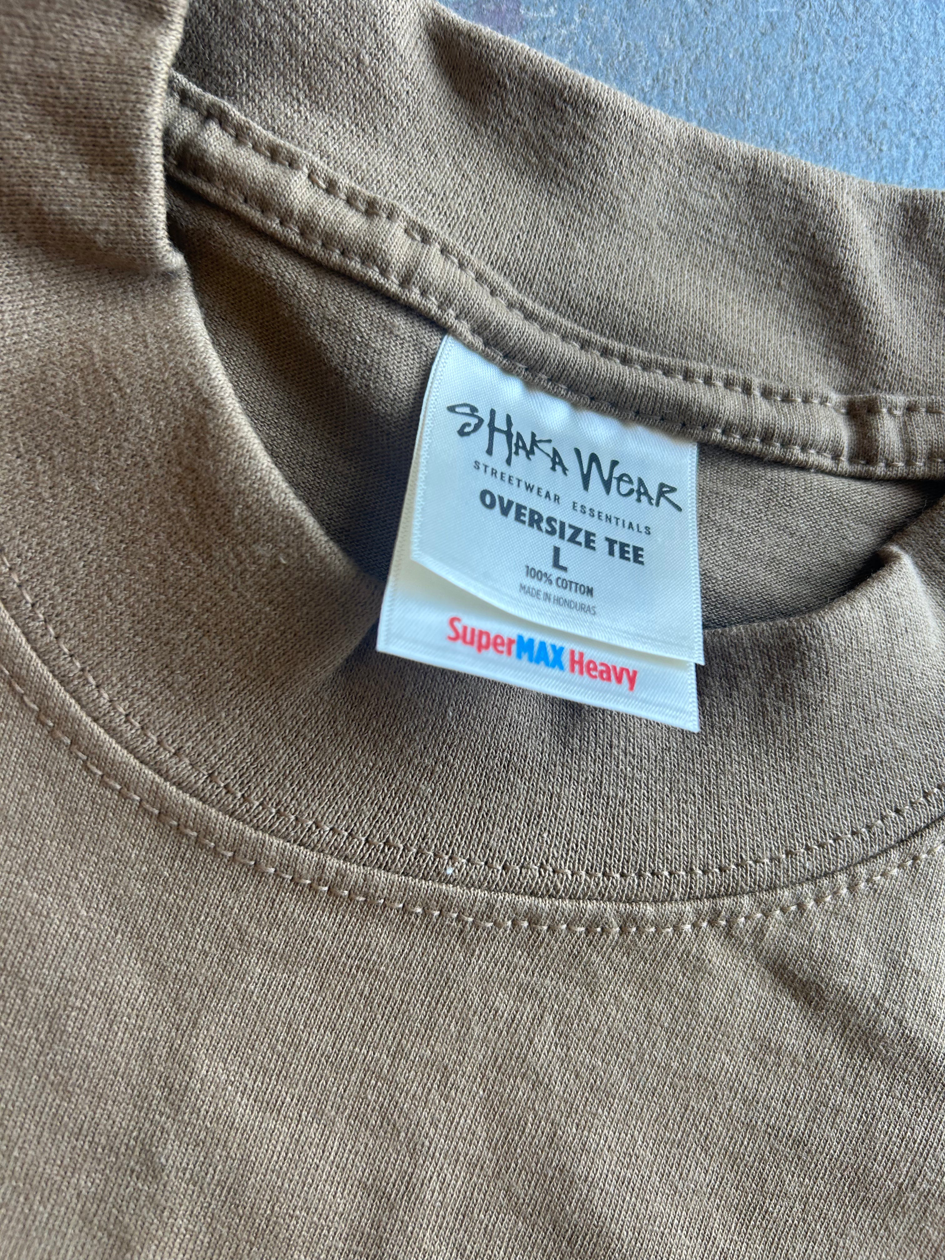 Close-up of a taupe brown oversized cotton t-shirt showing the inner neck label which reads 'SHAWA WEAR' and 'SuperMAX Heavy'.