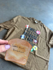 A hand with lavender nail polish holds a novelty slice of toasted bread in the foreground, with a brown graphic T-shirt laid out in the background on a gray floor. The shirt has colorful circular pins and black text reading 'WILD TIECED BECAUSE I AM A.