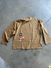 A tan long-sleeve shirt with three circular billiard-themed patches (black 8, red 1, and white 15) on the front, laid flat on a gray concrete floor.