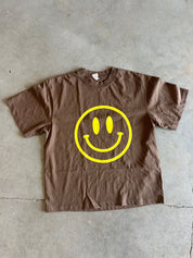 A brown short-sleeve crewneck t-shirt with a large yellow smiley face graphic printed on the front, laid flat on a gray concrete floor.
