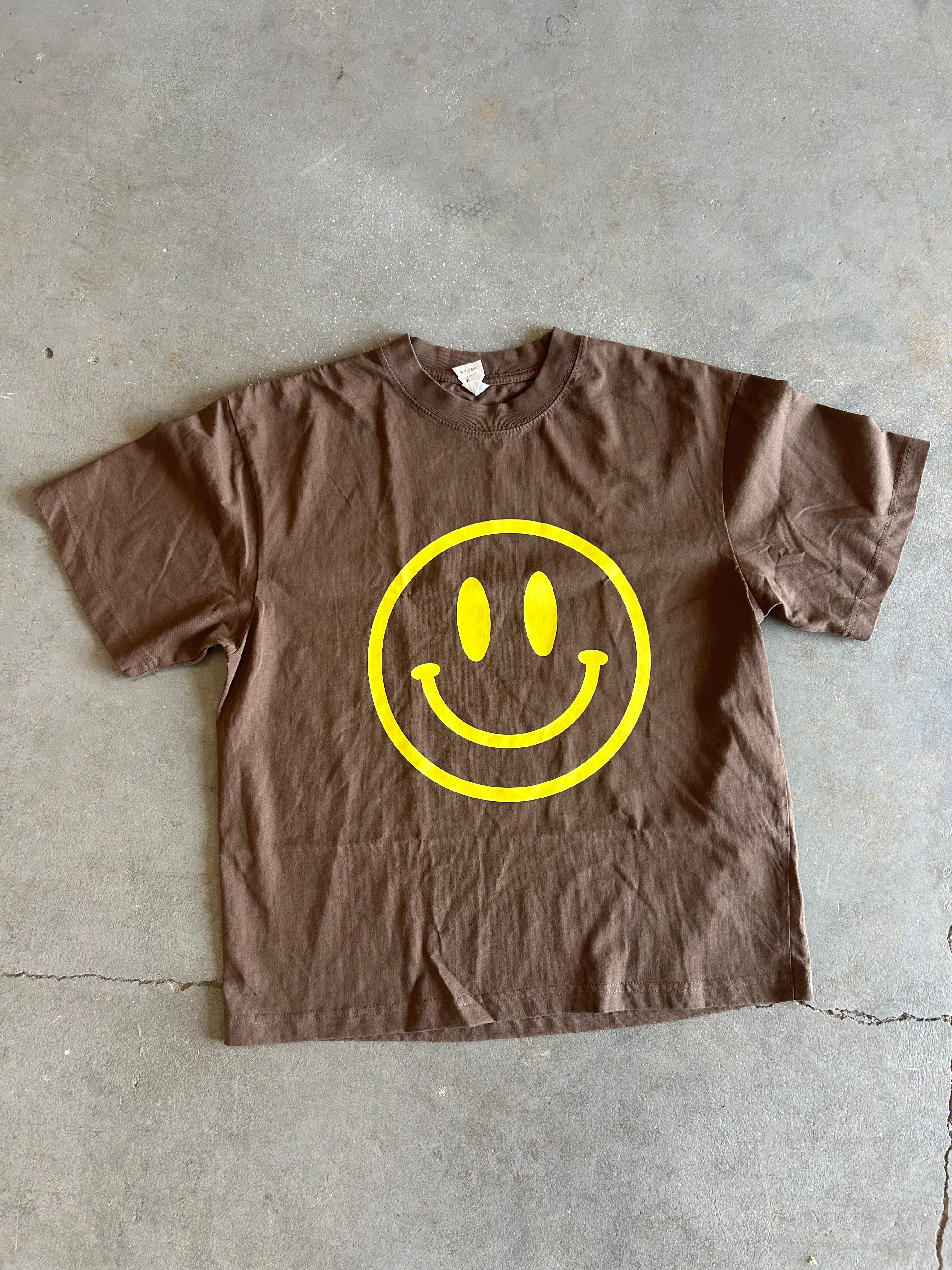 A brown short-sleeve crewneck t-shirt with a large yellow smiley face graphic printed on the front, laid flat on a gray concrete floor.