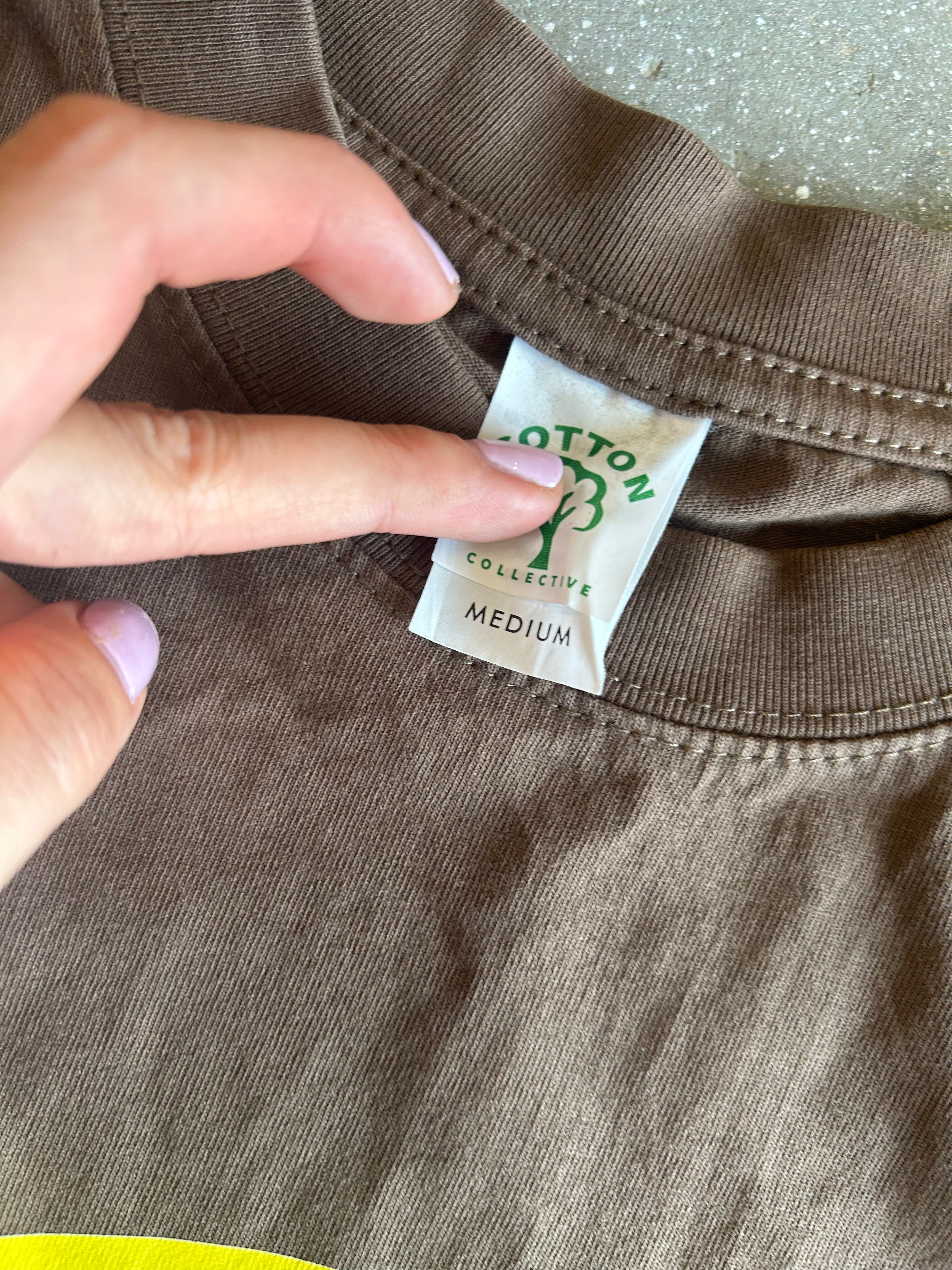 A close-up of a brown cotton t-shirt showing the inner neck label which reads 'COTTON COLLECTIVE' and 'MEDIUM'. A hand is pointing at the label.