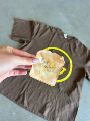 A hand holds a toasted slice of bread with handwritten text over a brown T-shirt laid on a gray concrete floor. The shirt has a yellow circular graphic and the word 'ENTER' printed on it.