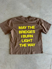 A brown short-sleeve t-shirt laid flat on a gray concrete floor, featuring the text 'MAY THE BRIDGES I BURN LIGHT THE WAY' printed in bold yellow capital letters on the front.