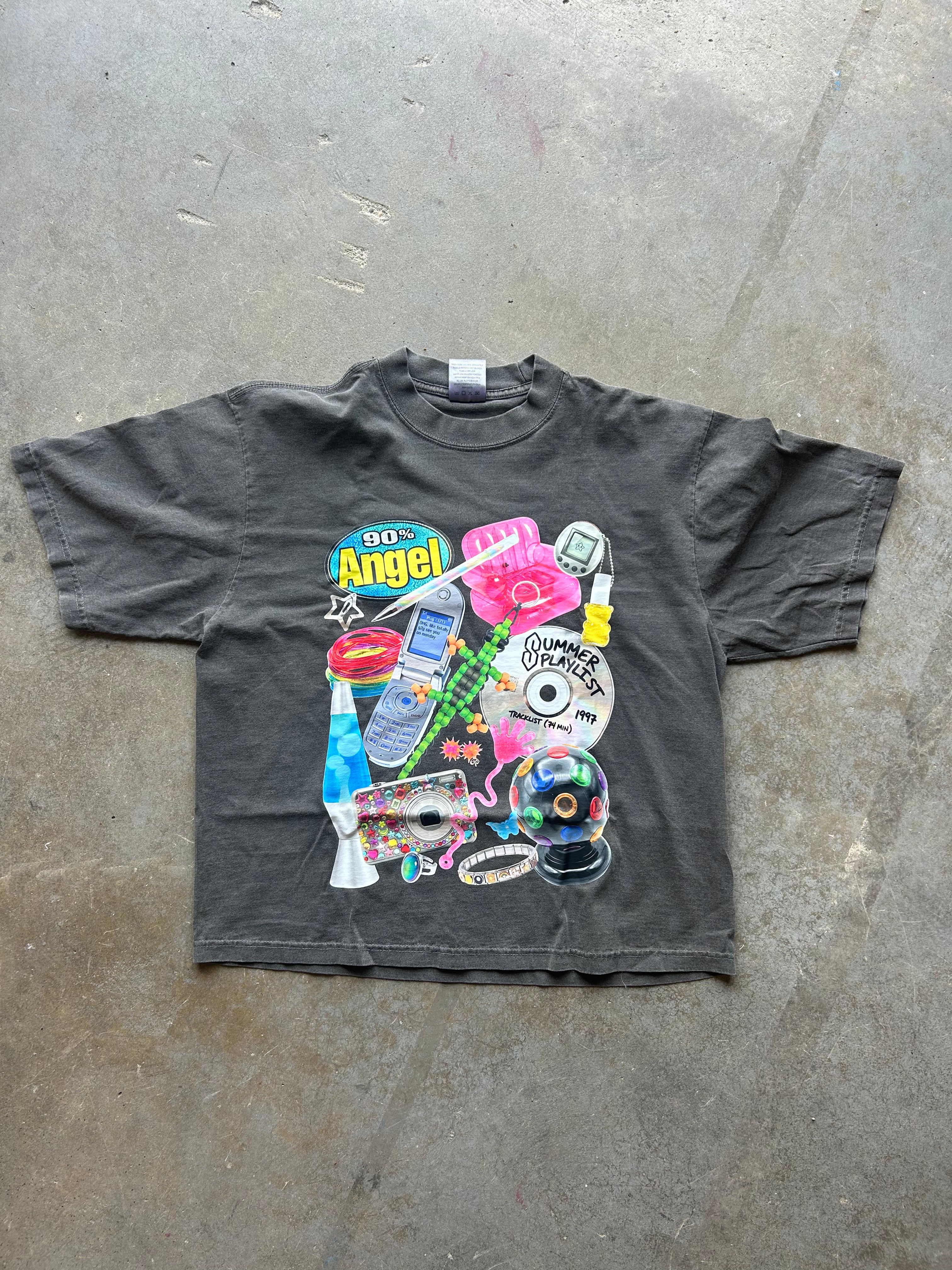 A dark gray graphic t-shirt laid flat on a concrete floor. The shirt features a large, colorful collage print on the front with retro pop culture icons, including a pink camera, a green cassette tape, a CD, a flip phone, and a 900 Angel logo.