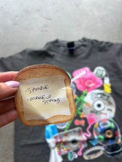 A hand holds a small, toast-shaped sample card with handwritten text reading '- SAMPLE - MAKEUP STING' in front of a black graphic t-shirt. The background is a concrete floor.