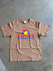 A tan short-sleeve t-shirt laid flat on a gray concrete floor, featuring a colorful graphic on the chest with the word 'MOTHER' in red and blue stripes and red, yellow, and blue circular shapes above it.