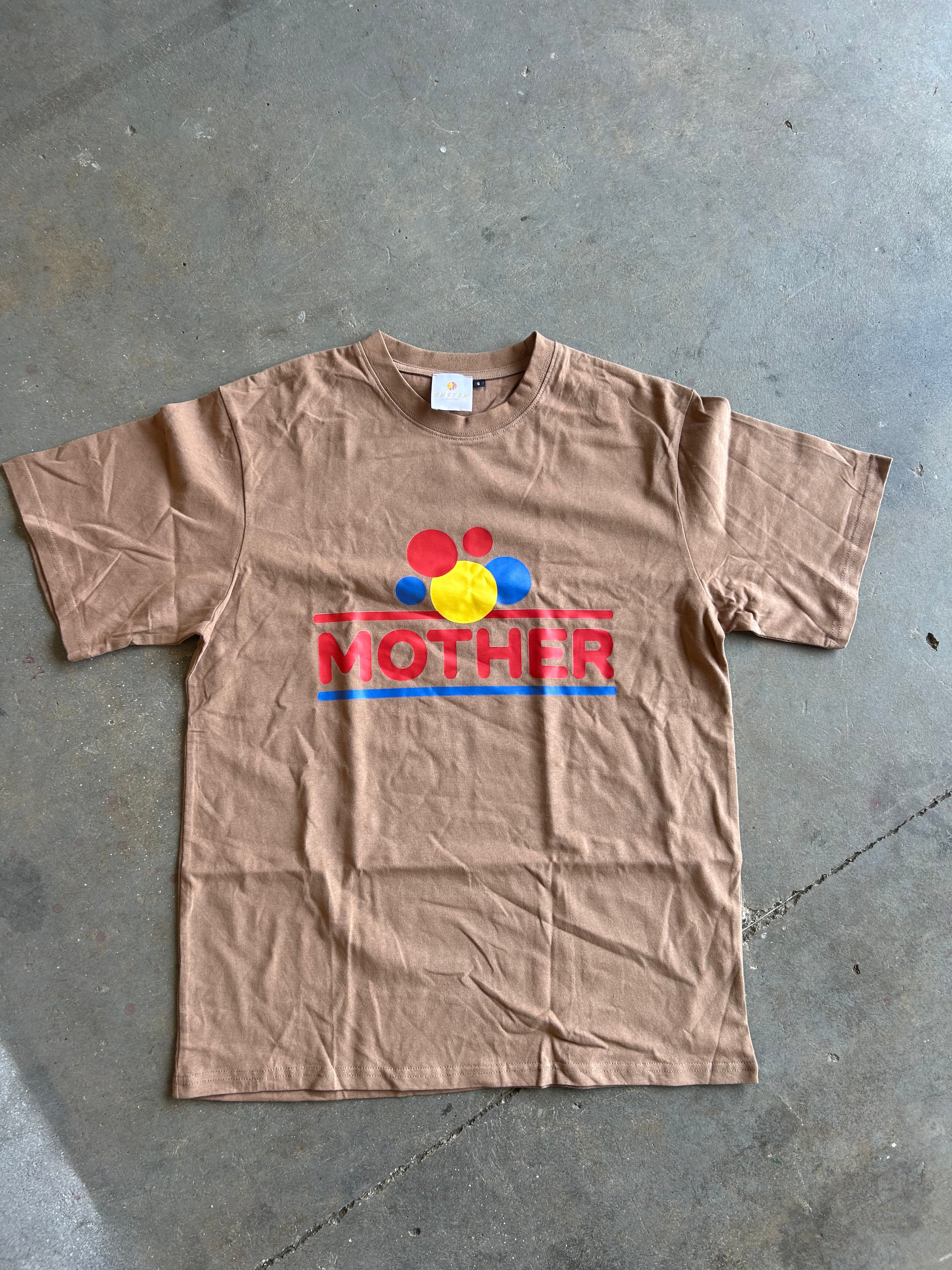 A tan short-sleeve t-shirt laid flat on a gray concrete floor, featuring a colorful graphic on the chest with the word 'MOTHER' in red and blue stripes and red, yellow, and blue circular shapes above it.
