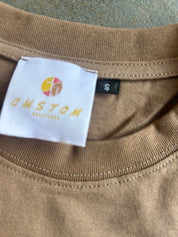 Close-up of a brown crewneck t-shirt showing a white custom label with a yellow and red logo and the text 'CUSTOM BEAUTY' alongside a black size tag marked 'S'.