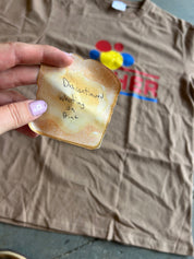 A hand with lavender nail polish holds a novelty toast-shaped item with handwritten text reading 'Discontinued Whipping on Print' in front of a wrinkled brown t-shirt with a colorful graphic.