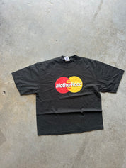 A black short-sleeve t-shirt laid flat on a gray concrete floor, featuring a parody graphic of the Mastercard logo with the word 'MotherHood' printed in white across the red and yellow circles.