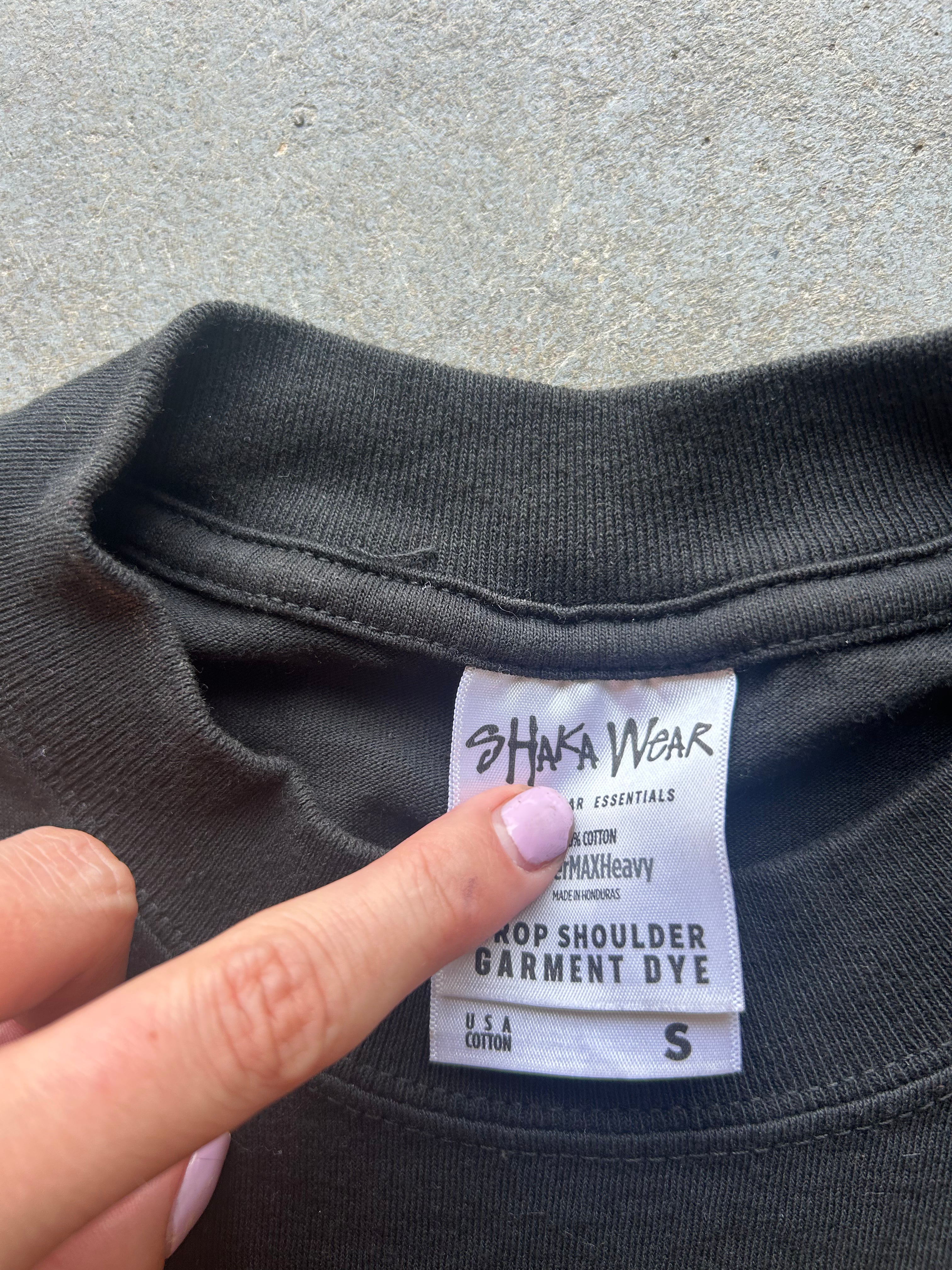 Close-up of a black sweatshirt neck label being pointed at by a finger. The visible brand on the label is Shaka Wear, and the text reads 'Shop Shoulder Garment Dye' with size S.