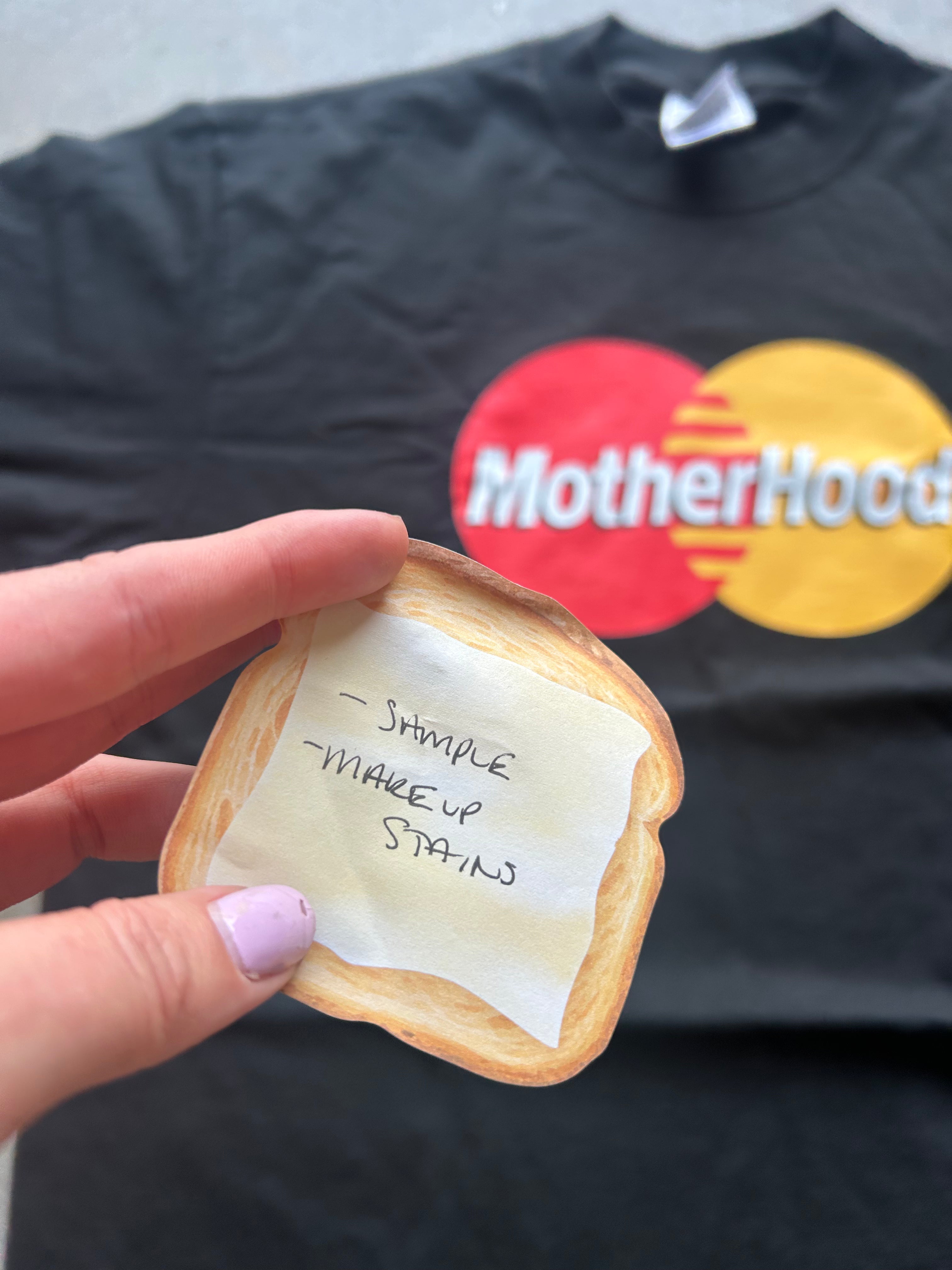 A hand holds a novelty toast-shaped item with handwritten text in front of a black t-shirt featuring a red and yellow Mastercard logo and the text 'MotherHood'.