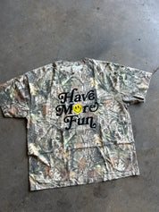 A short-sleeve camouflage t-shirt with the text 'Have More Fun' in black script and a yellow smiley face graphic, laid flat on a gray concrete floor.