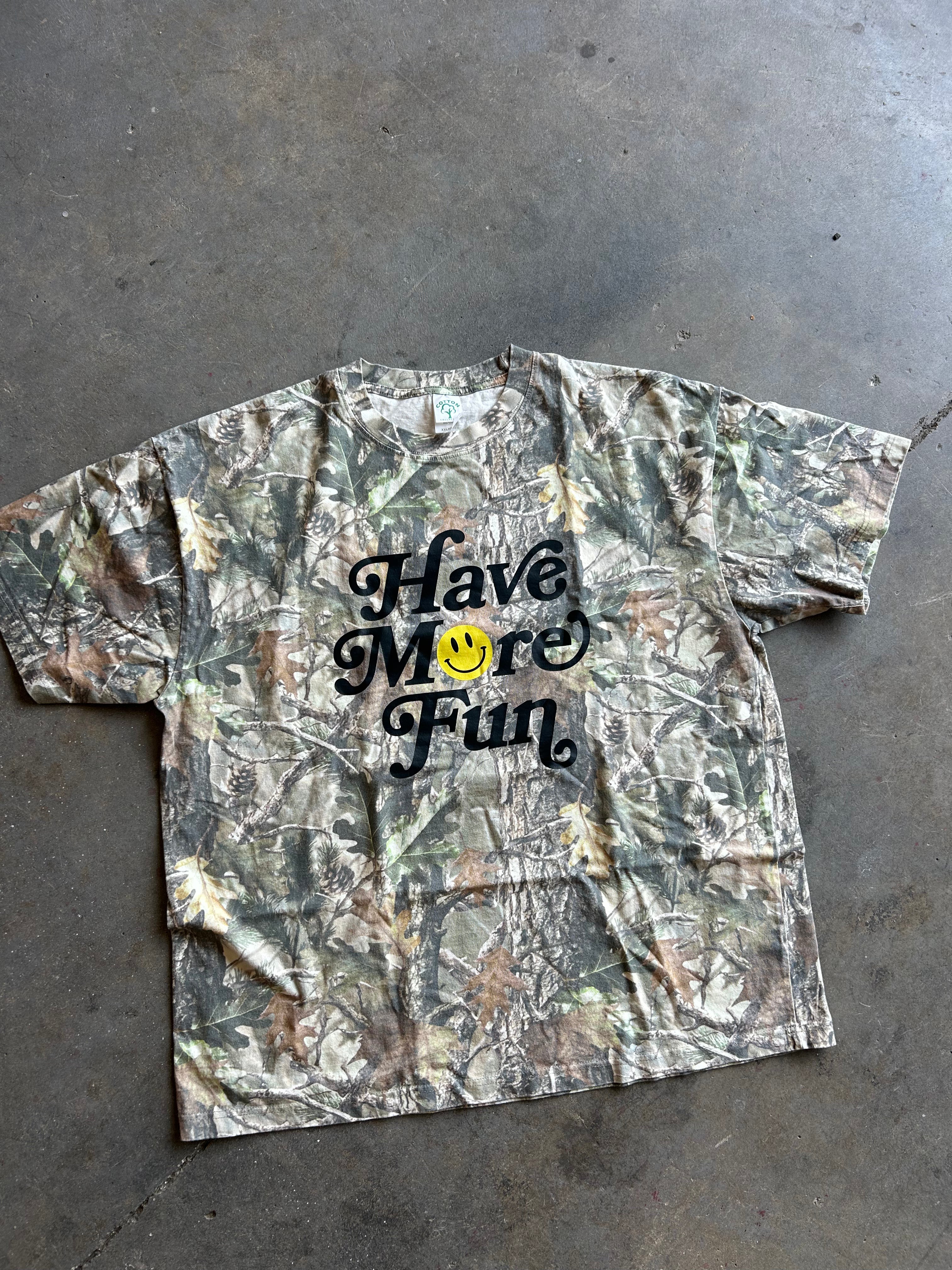 A short-sleeve camouflage t-shirt with the text 'Have More Fun' in black script and a yellow smiley face graphic, laid flat on a gray concrete floor.