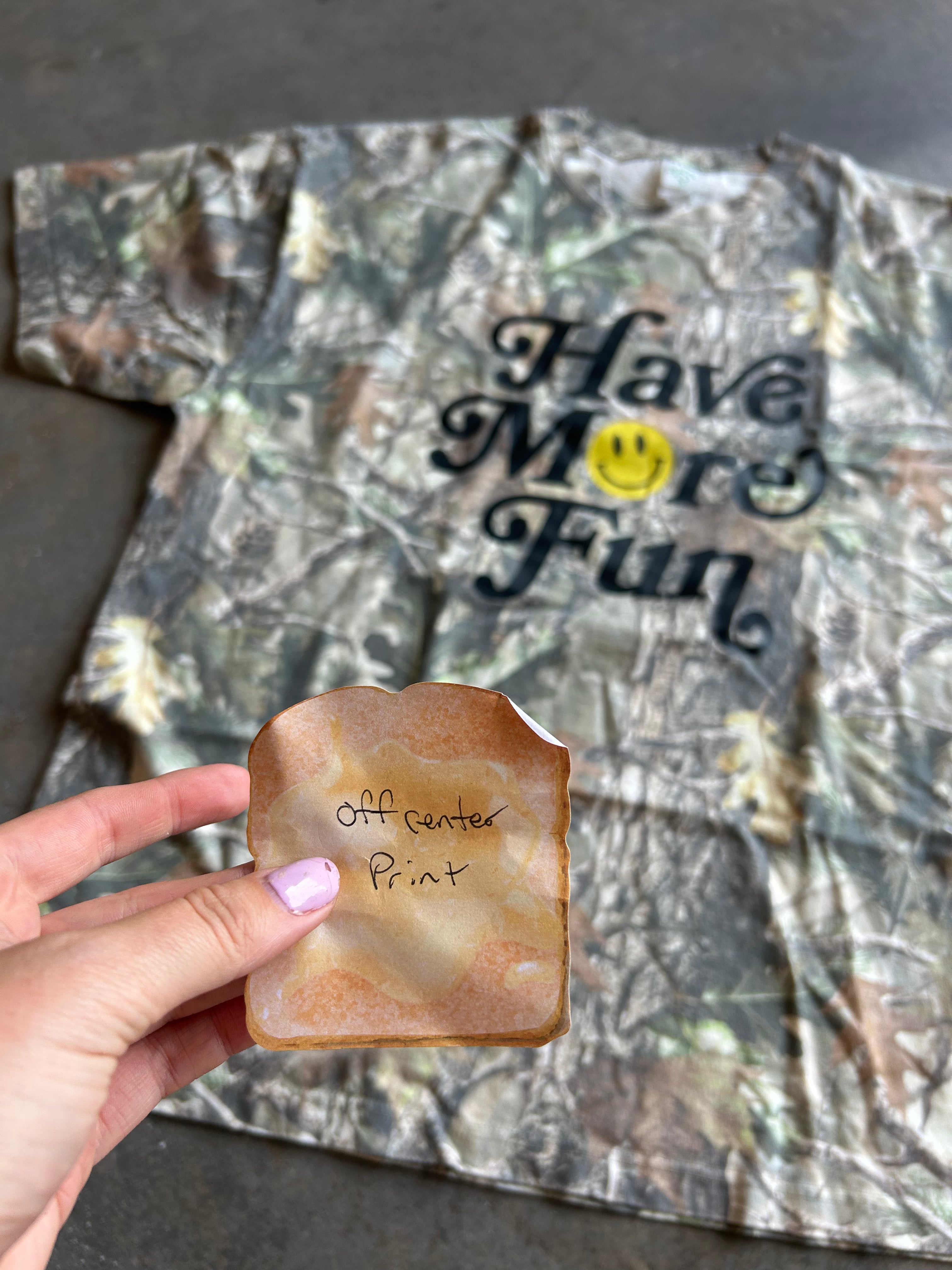 A hand holds a small, irregularly shaped tan and brown piece of material in front of a camouflage-print T-shirt with the text 'Have More Fun' and a yellow smiley face. The background is a real surface, and a hand is visible, making the subject type Lifestyle.