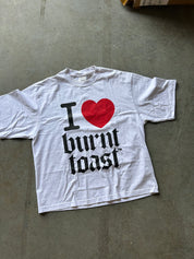 A white short-sleeve graphic T-shirt laid flat on a gray concrete floor, featuring a red heart and the text 'I [heart] burnt toast' in black gothic lettering.