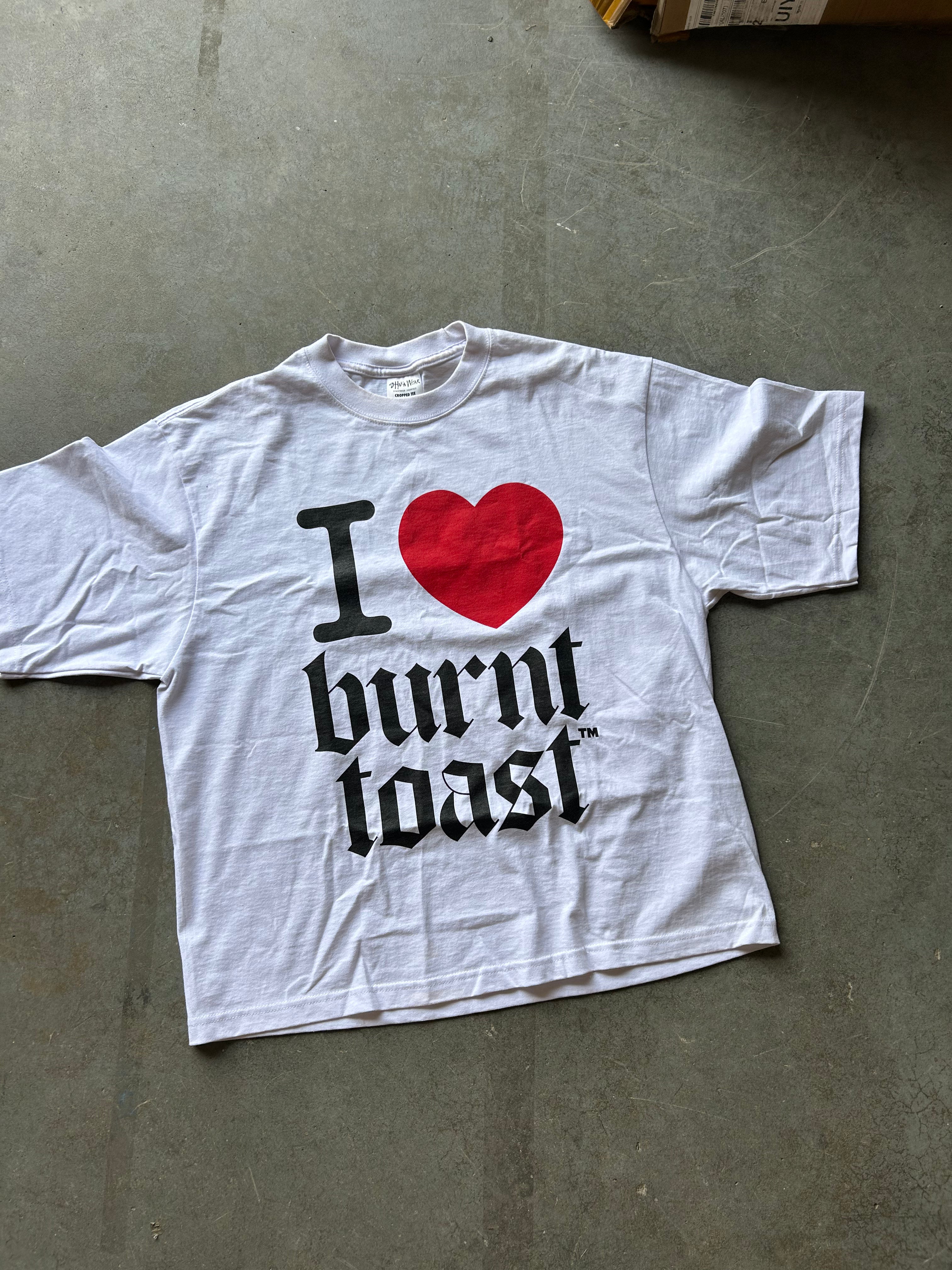 A white short-sleeve graphic T-shirt laid flat on a gray concrete floor, featuring a red heart and the text 'I [heart] burnt toast' in black gothic lettering.
