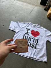 A white graphic T-shirt laid on a concrete floor with a hand holding a piece of burnt toast in front of it. The shirt has a red heart and black gothic text reading 'I [heart] burnt toast'.