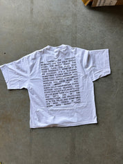 A white short-sleeve T-shirt laid flat on a gray concrete floor, featuring a black printed poem on the front.