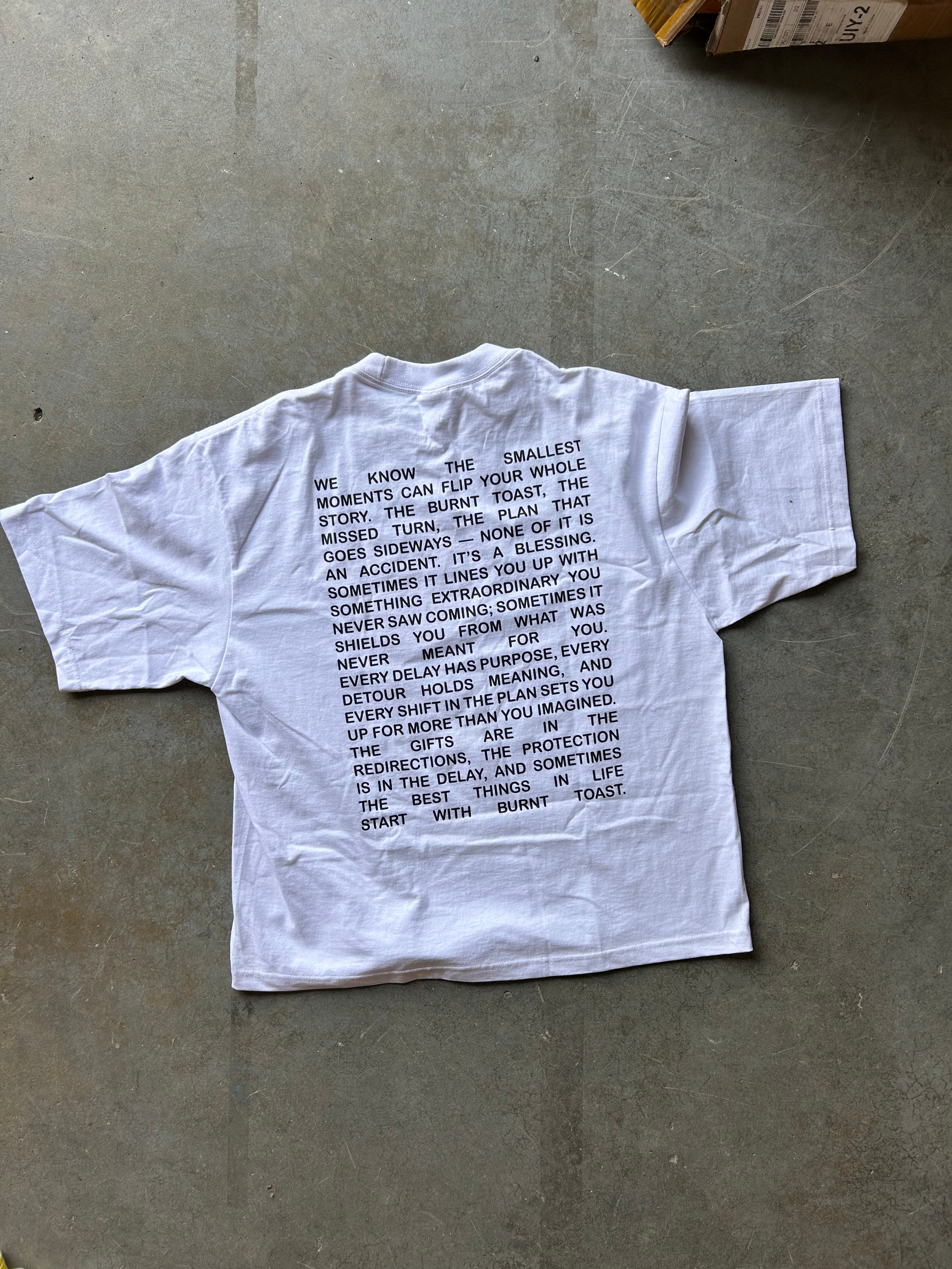 A white short-sleeve T-shirt laid flat on a gray concrete floor, featuring a black printed poem on the front.