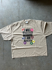 A beige short-sleeve t-shirt laid flat on a gray concrete floor, featuring a graphic print of colorful billiard balls surrounding the text 'I WILL SUCCEED BECAUSE I AM Insane'.