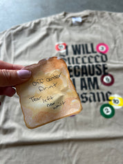 A beige short-sleeve t-shirt laid flat on a gray concrete floor, featuring a graphic print of colorful billiard balls surrounding the text 'I WILL SUCCEED BECAUSE I AM Insane'.