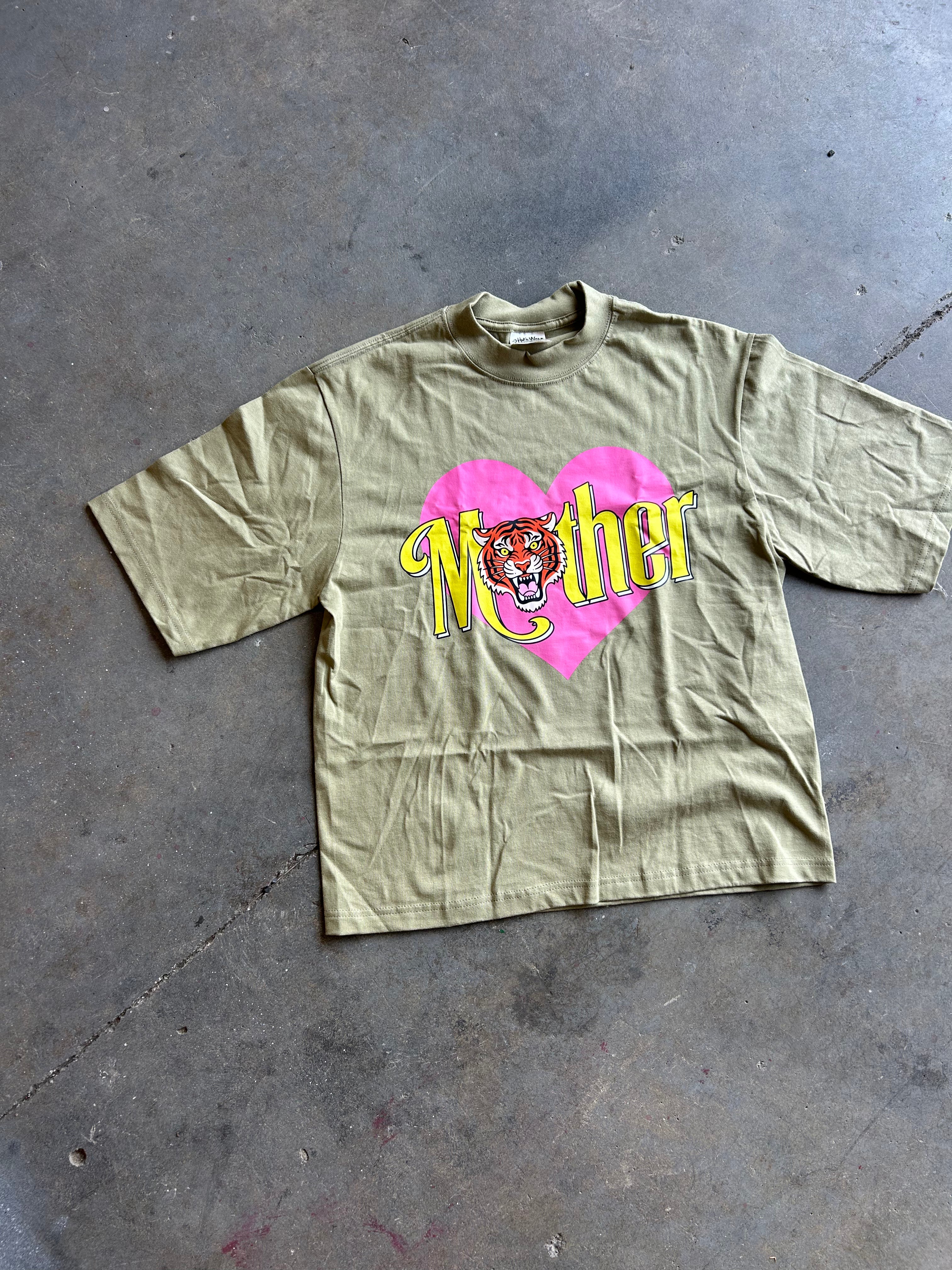 An olive green short-sleeve t-shirt with a graphic print of a pink heart, yellow 'Mother' text, and a tiger face, laid flat on a gray concrete floor.