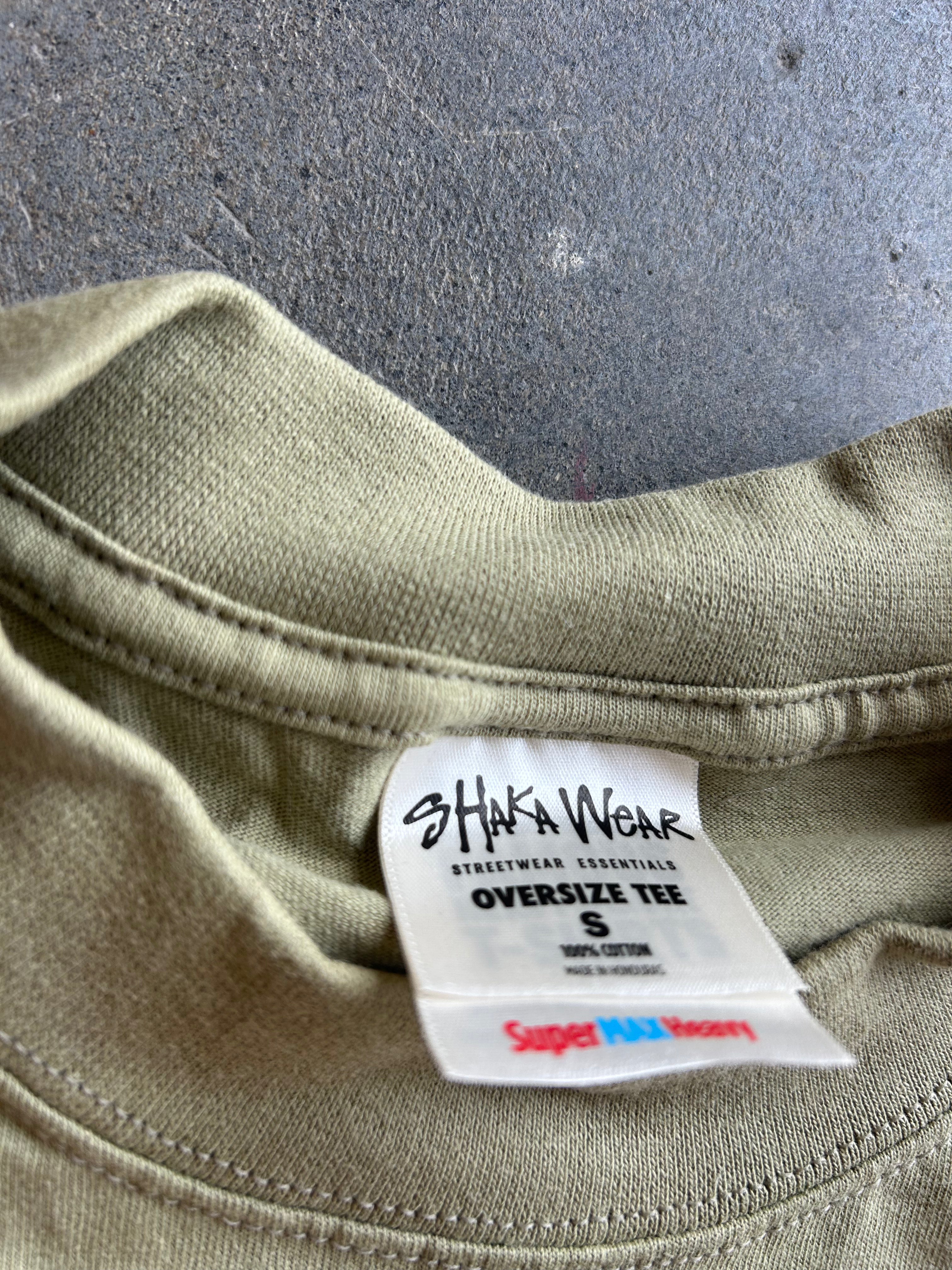Close-up of the inside collar of an oversized olive green t-shirt, showing a white brand label for 'Shaka Wear' and a 'Super Pile Cotton' tag.