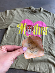 An olive green t-shirt with a pink heart and yellow 'Mother' tiger graphic, with a hand holding a piece of toast marked 'Off Center Print' in front of it.