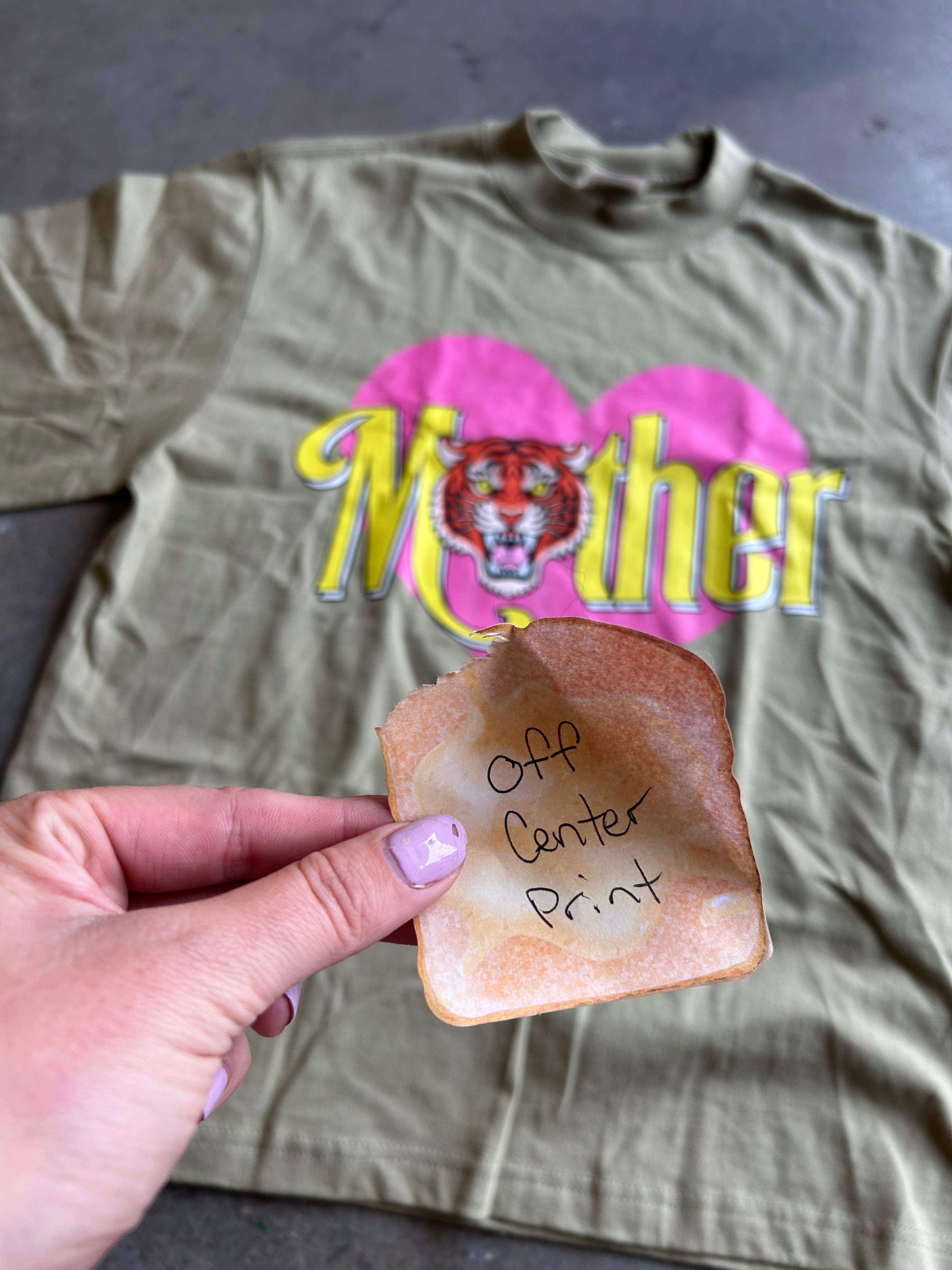 An olive green t-shirt with a pink heart and yellow 'Mother' tiger graphic, with a hand holding a piece of toast marked 'Off Center Print' in front of it.
