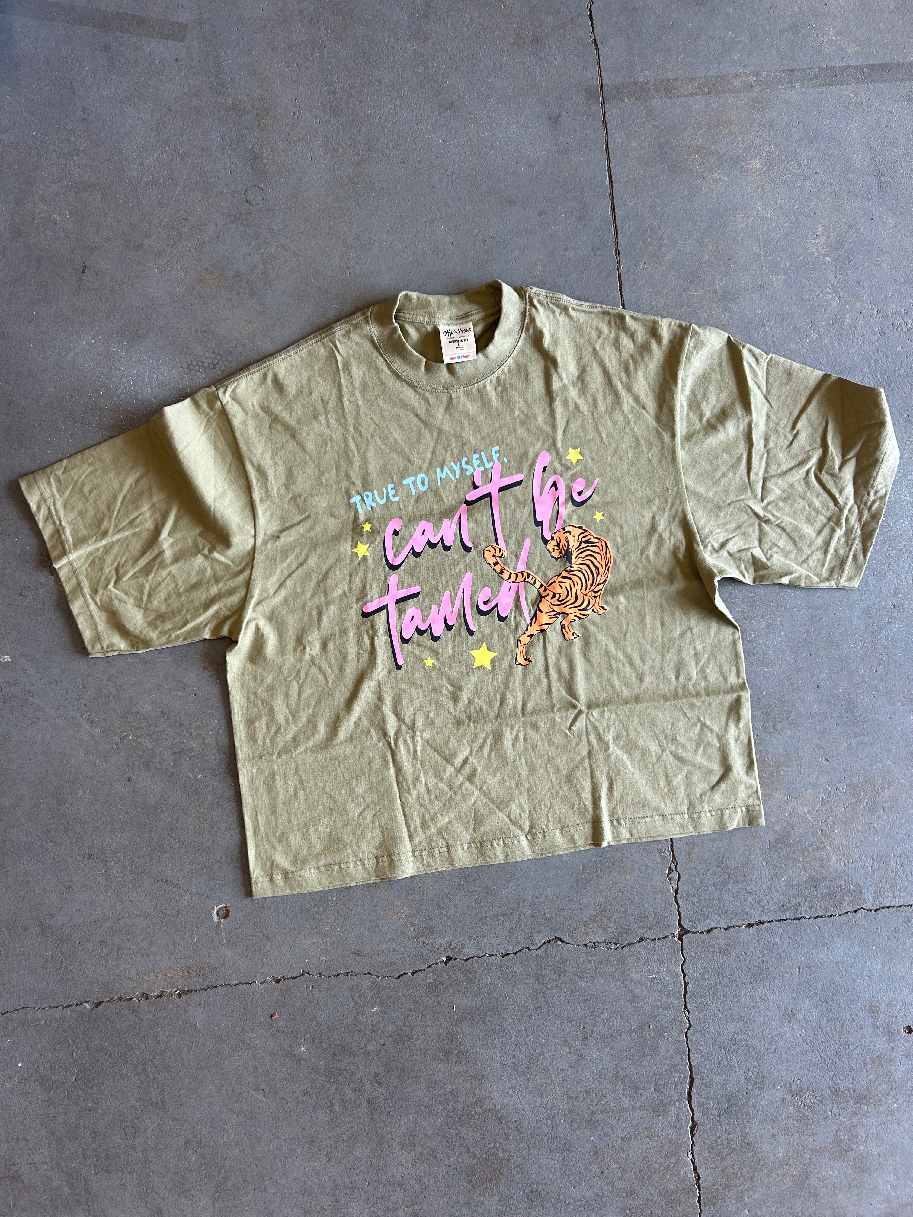 A sage green graphic t-shirt laid flat on a gray concrete floor. The shirt features a colorful graphic on the front with the text 'TRULY TO MYSELF can't be tamed' in pink and blue, accented with yellow stars and an orange tabby tiger illustration.