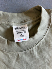 Close-up of an oversized crewneck t-shirt in a muted sage green color, showing the inner neck label for 'Shaka Wear'.