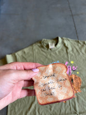 A hand holds a novelty toast-shaped tag with handwritten text over an olive green t-shirt laid flat on a gray tiled floor.