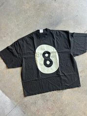 A black short-sleeve t-shirt laid flat on a concrete floor, featuring a large cream-colored oval graphic on the front with a black number 8 in the center.