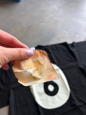 A hand with lavender nail polish holds a small, burnt beige paper tag with handwritten text over a black t-shirt featuring a white and black graphic.