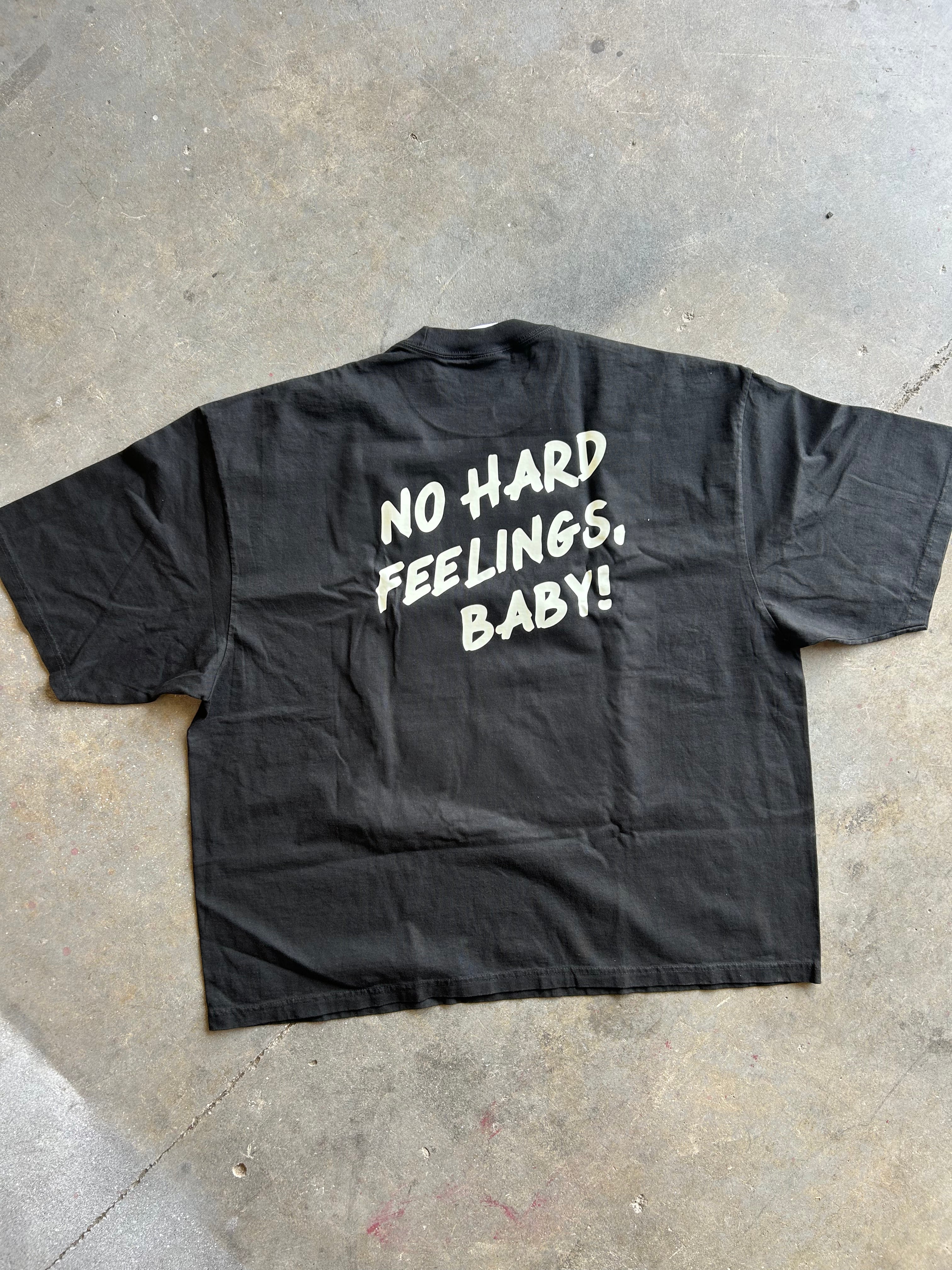 A black short-sleeve t-shirt laid flat on a concrete floor, showing the back with the phrase 'NO HARD FEELINGS, BABY!' printed in white hand-drawn style lettering.