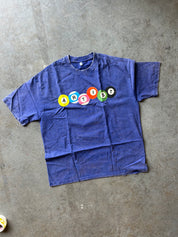 A faded blue short-sleeve t-shirt laid flat on a gray concrete floor, featuring the word 'ARTIST' spelled out in colorful circular letter stickers across the chest.