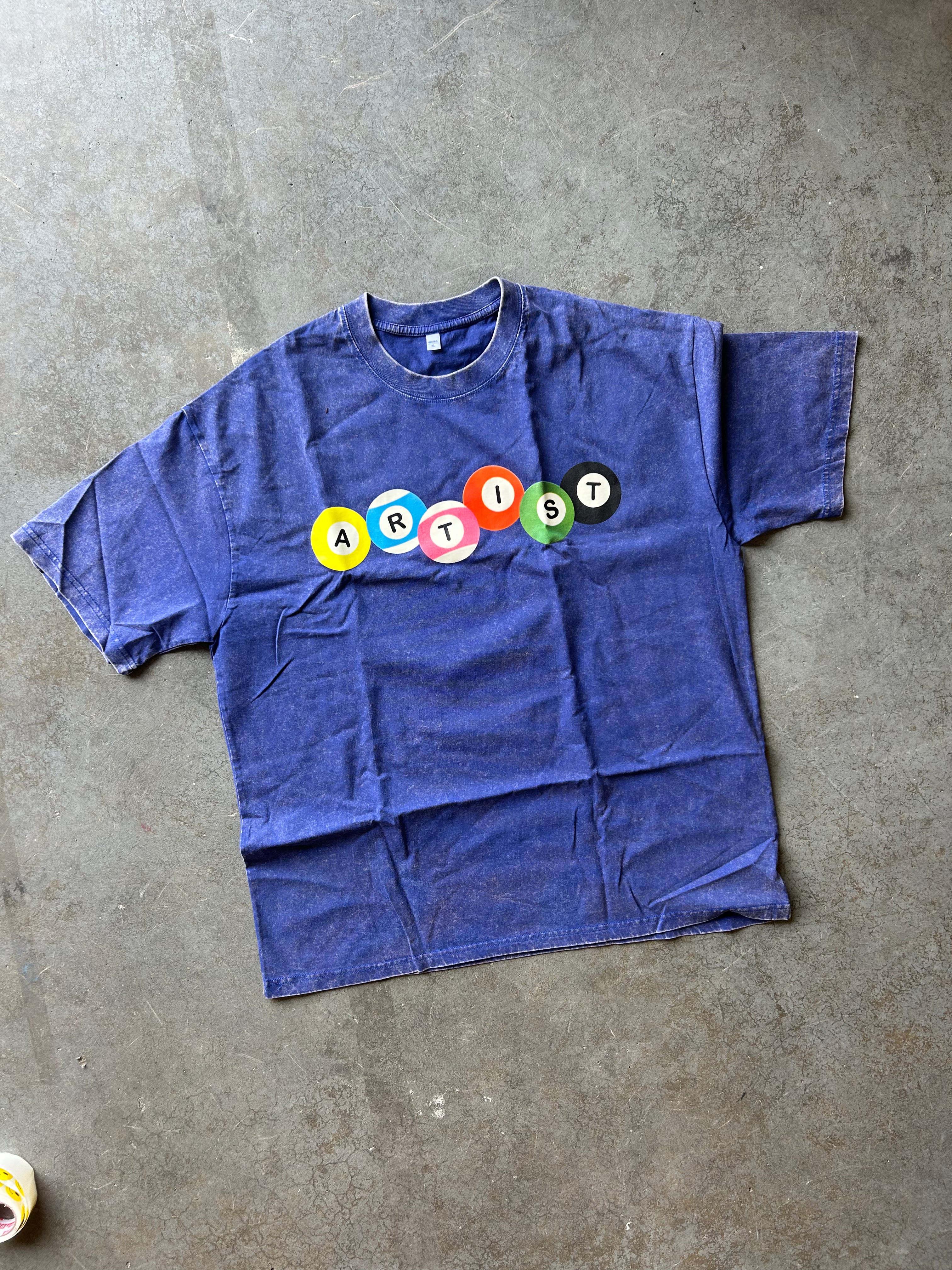 A faded blue short-sleeve t-shirt laid flat on a gray concrete floor, featuring the word 'ARTIST' spelled out in colorful circular letter stickers across the chest.