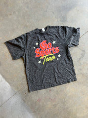 A dark gray heathered graphic t-shirt laid flat on a concrete floor. The shirt features a red and yellow 'Go Sports Team' graphic with white stars.
