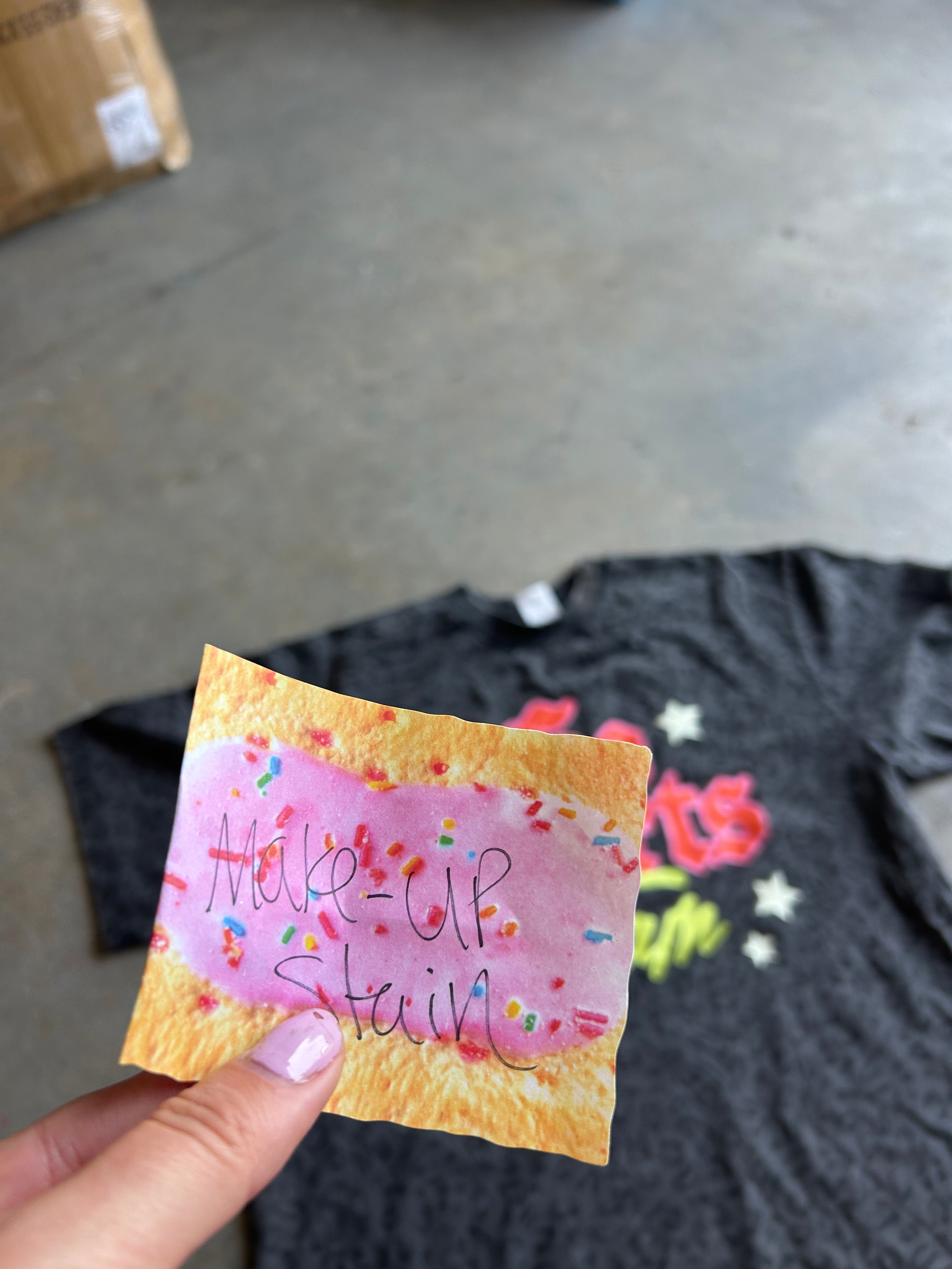 A hand with lavender nail polish holds a small printed card featuring a pink frosted pastry with colorful sprinkles and handwritten text. In the blurred background, a black graphic T-shirt is laid out on a concrete floor.