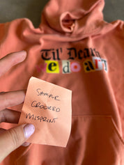 A close-up of a peach-colored hoodie with a colorful 'we do art' graphic on the chest, held by a hand holding a sticky note that reads 'Sample Crooked Misprint'.