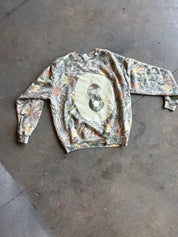 A long-sleeve crewneck sweatshirt with a Realtree camouflage pattern and a large beige oval graphic on the chest featuring a creme 8 ball, laid flat on a concrete floor.