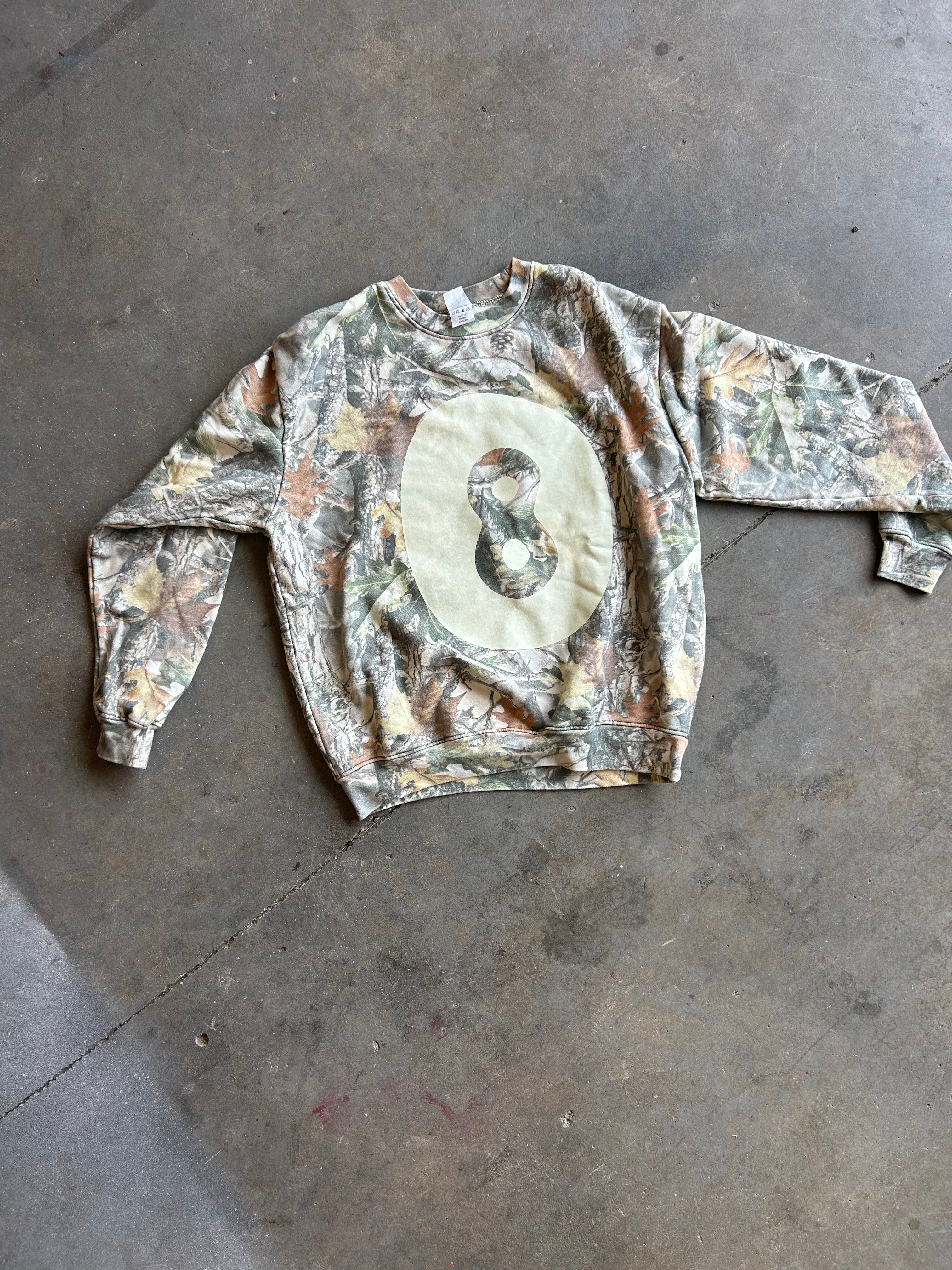 A long-sleeve crewneck sweatshirt with a Realtree camouflage pattern and a large beige oval graphic on the chest featuring a creme 8 ball, laid flat on a concrete floor.
