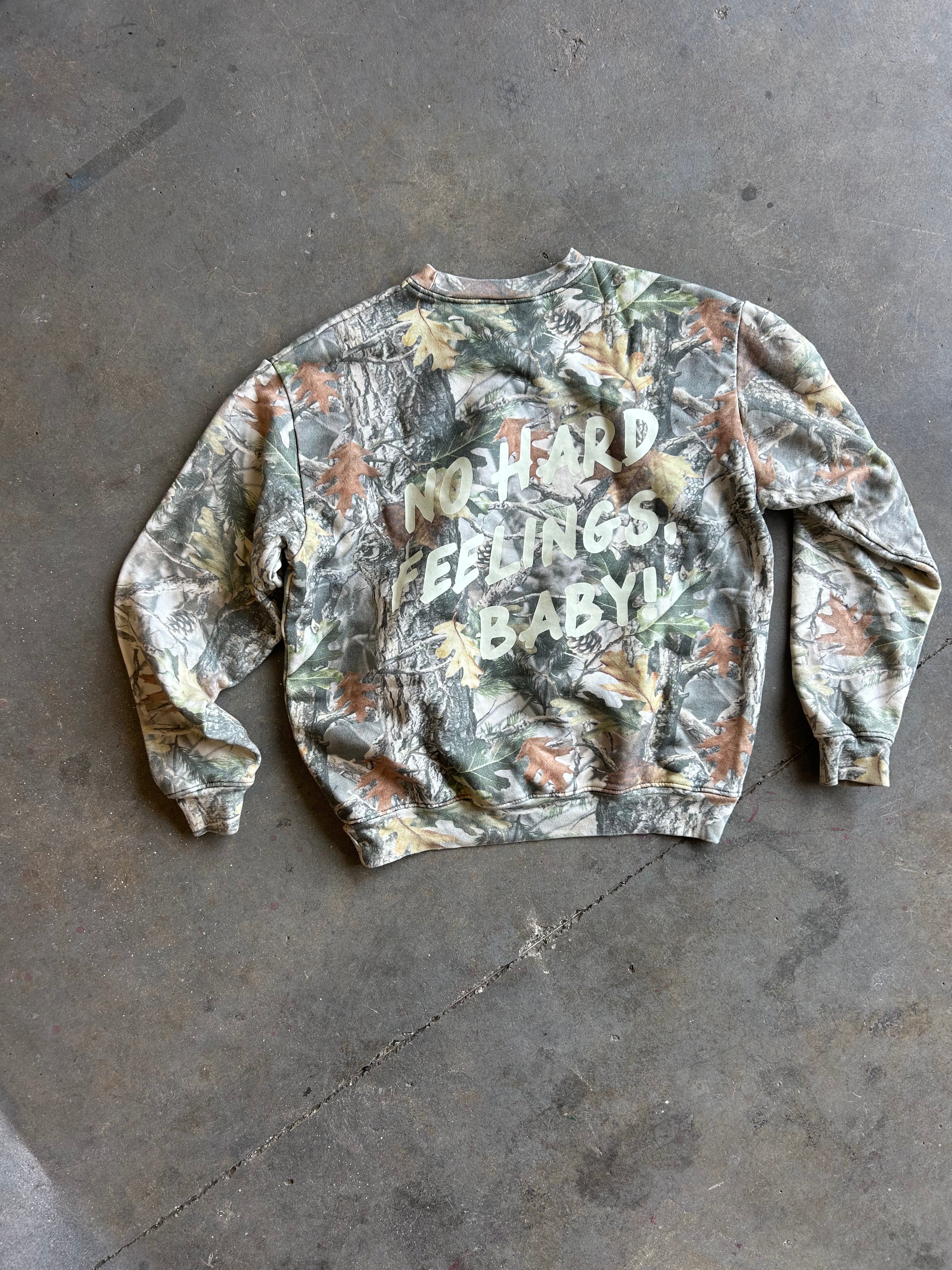A camouflage crewneck sweatshirt with the text 'I'NO HARD FEELINGS BABY!' printed in white across the chest, laid flat on a gray concrete floor.