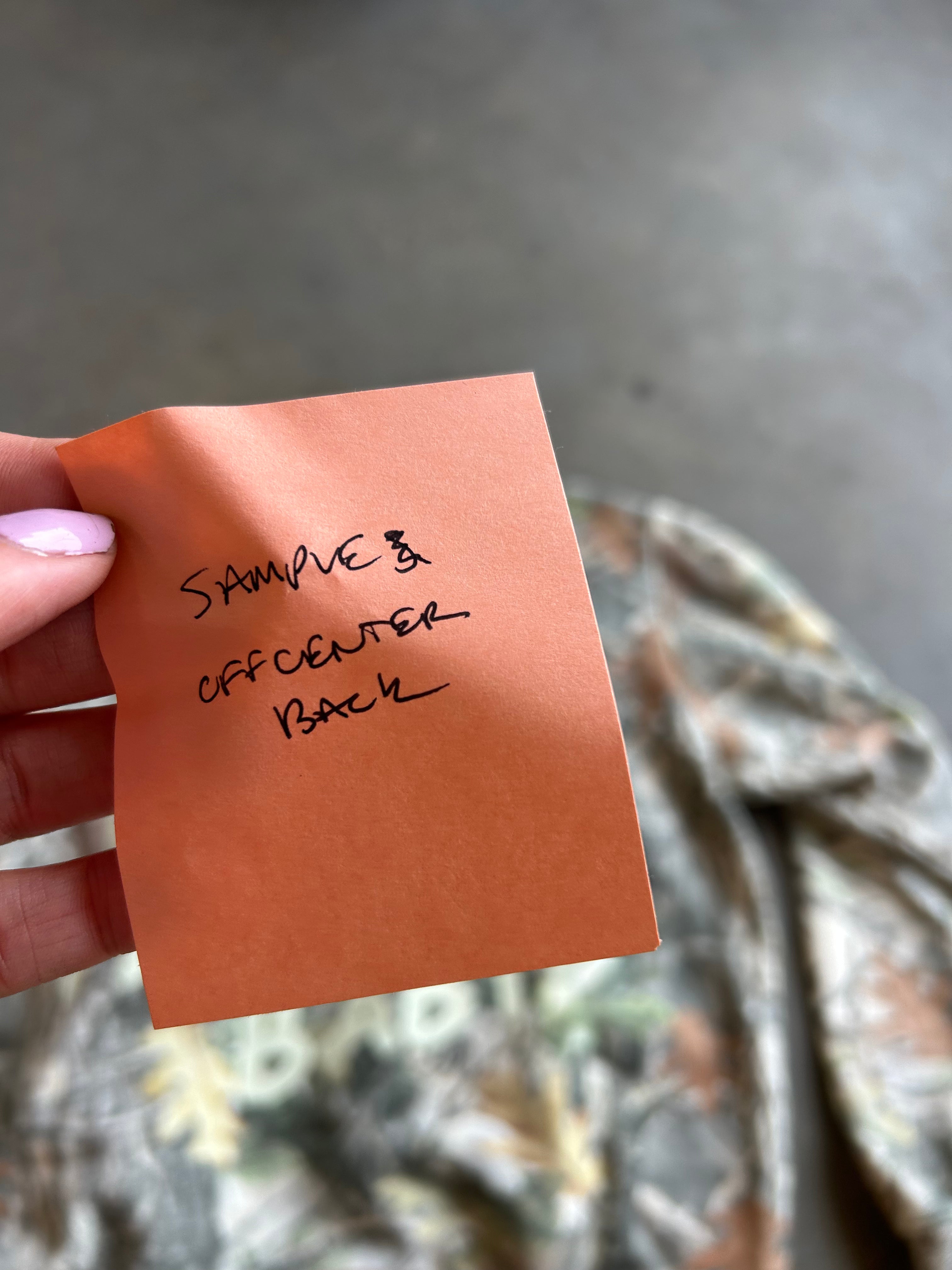 A hand with pink nail polish holds a small orange paper note with handwritten text reading 'SAMPLE OFF CENTER BACK' over a blurred camouflage-patterned fabric and gray concrete floor.