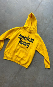 A bright yellow pullover hoodie with 'American Honey' printed in black across the chest, laid flat on a gray concrete floor.