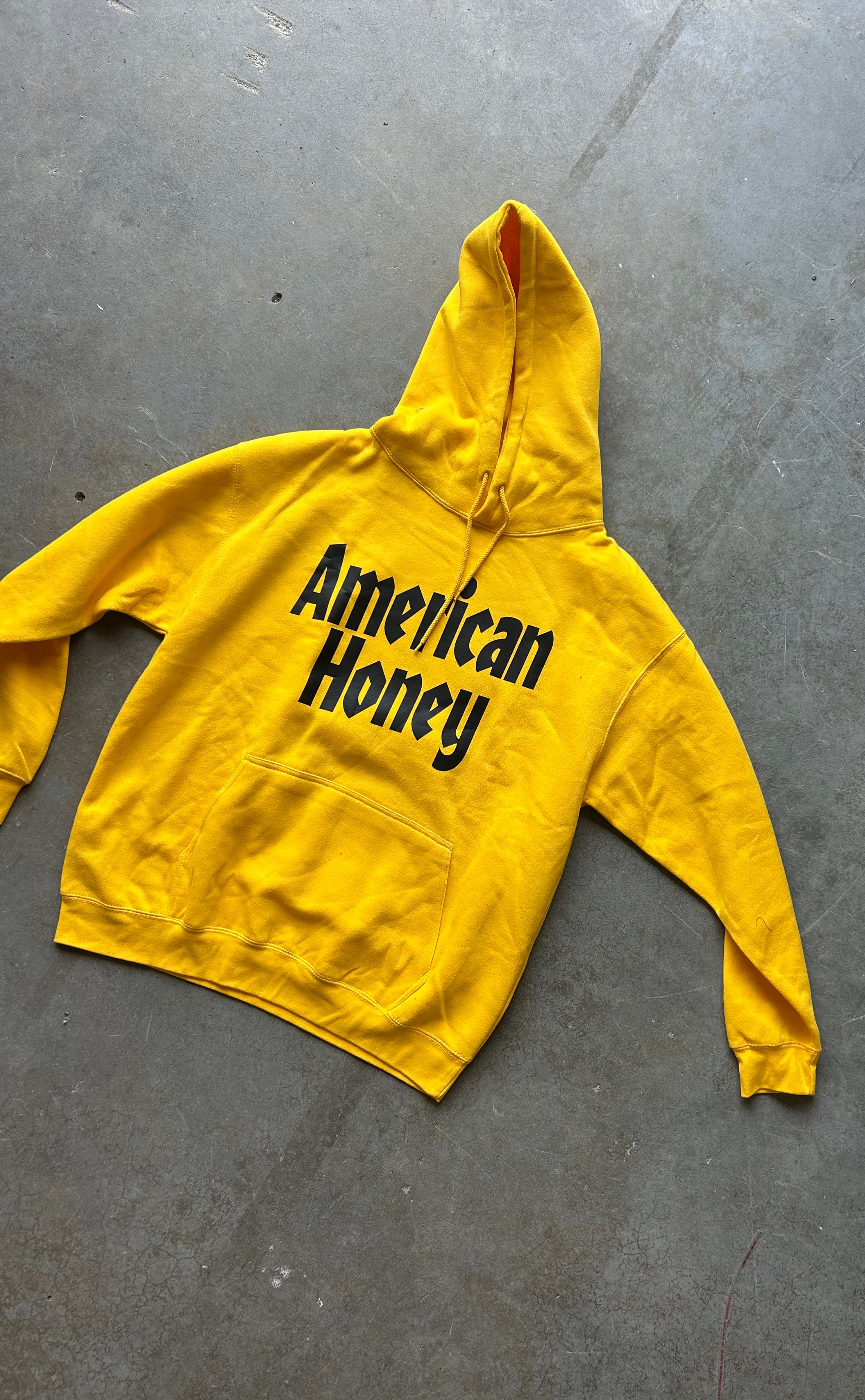 A bright yellow pullover hoodie with 'American Honey' printed in black across the chest, laid flat on a gray concrete floor.