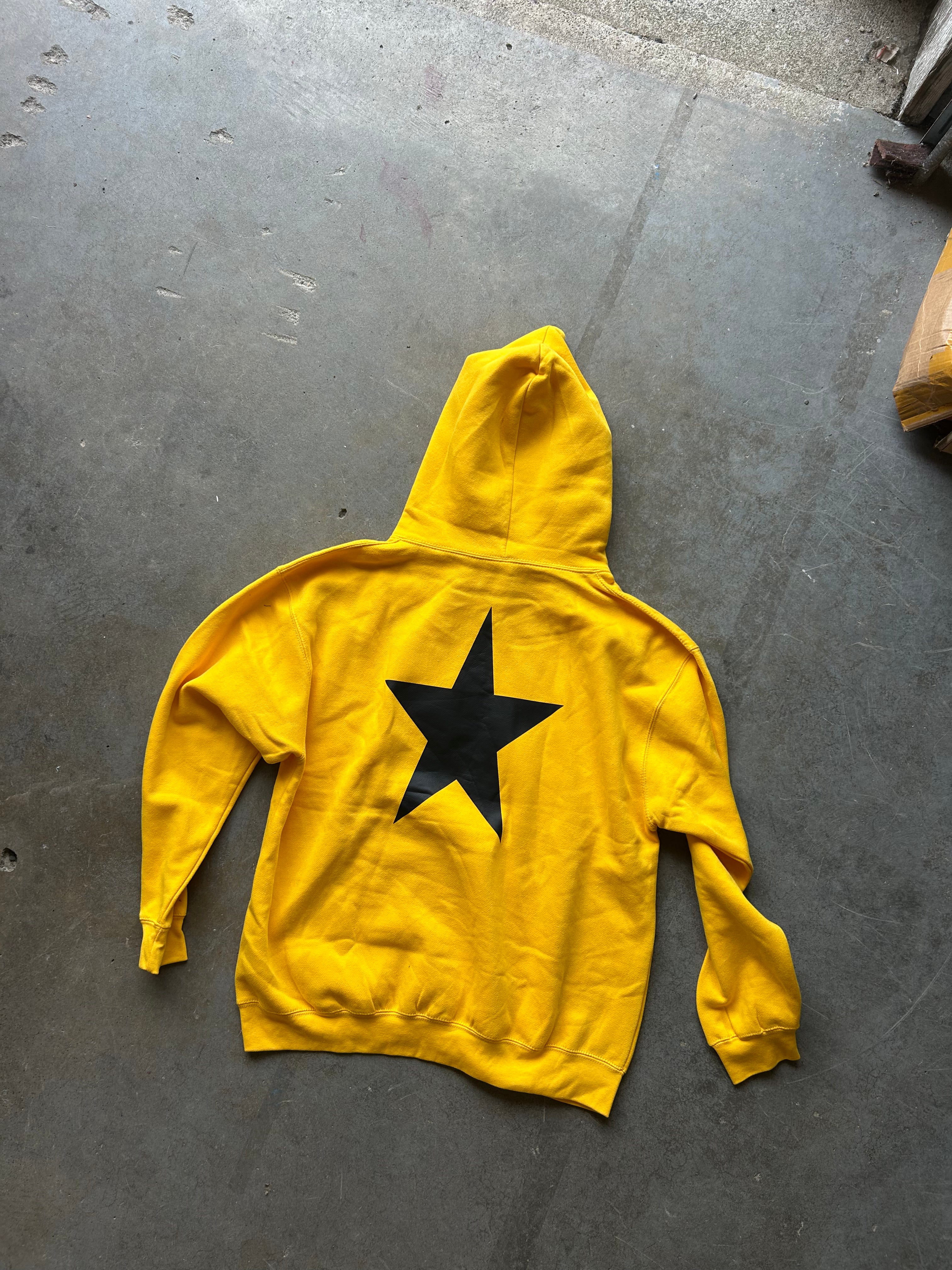 A bright yellow hoodie laid flat on a gray concrete floor, featuring a large black star graphic printed on the back.